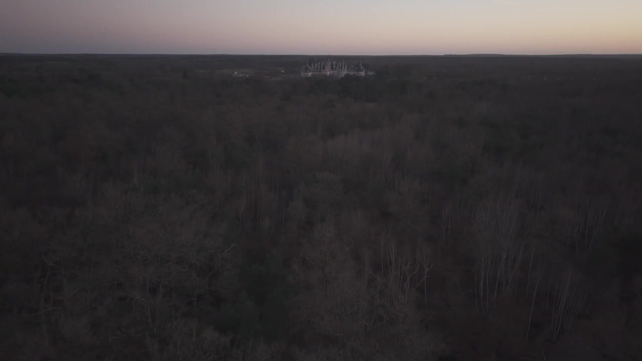 Uma vista aérea passa sobre planícies e campos abertos escurecendo ao anoitecer, perto do Castelo de Chambord, França.