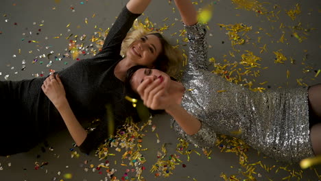 two female friends lying on the floor full of golden confetti after a glamorous new years eve party, laughing and dancing