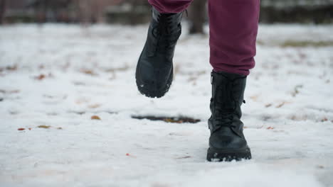 close-up of legs wearing black boots as person jumps with one leg on snow-covered ground, snow slightly shifts underfoot, creating subtle motion