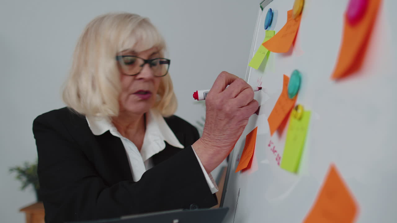 Mulher de negócios idosa treinadora oradora de conferência analisando infográficos desenhando no quadro branco do escritório