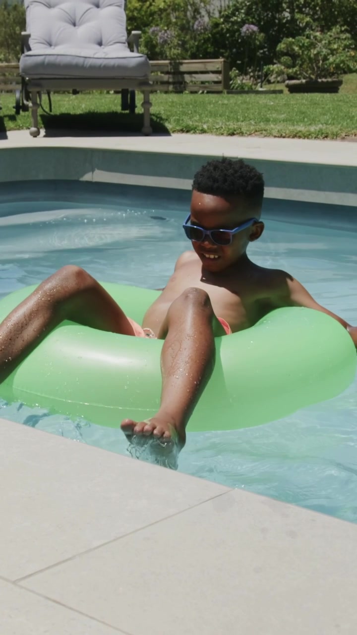 Vertical video: Shifting kicking kid in pink trunks, shades moving green ring to backyard pool edge