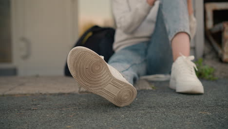 primer plano de la pierna del individuo sentado, centrándose en la suela de la zapatilla blanca, con la mano descansando en la pierna y la bolsa negra cerca, el entorno al aire libre casual hace hincapié en el estado de ánimo relajado y relajado