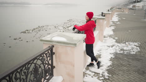 woman in red hoodie and black leggings walks toward iron fence and rests hands on decorative railing overlooking large calm water body, surrounded by snow-covered ground