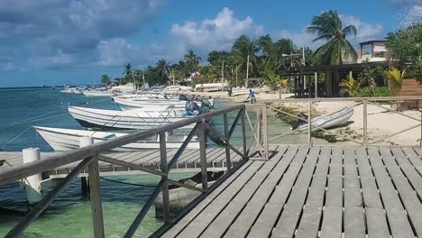 WOODEN-FISHING-PIER-WITH-WHITE-MOTOR-BOATS-MOORED-COAST,-PALMS-TREE-AND-SMALL-TROPICAL-VILLAGE