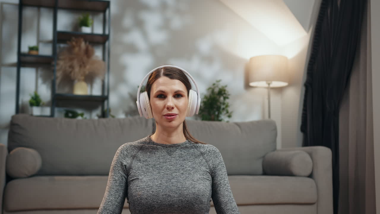 Pregnant woman sits in lotus pose wearing headphones eyes closed meditating peacefully at home