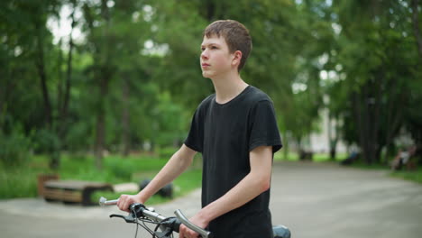 young boy in black top and ash pants is walking his bicycle along park pathway, the scene is surrounded by trees and greenery, with a blurred background of distant people and buildings