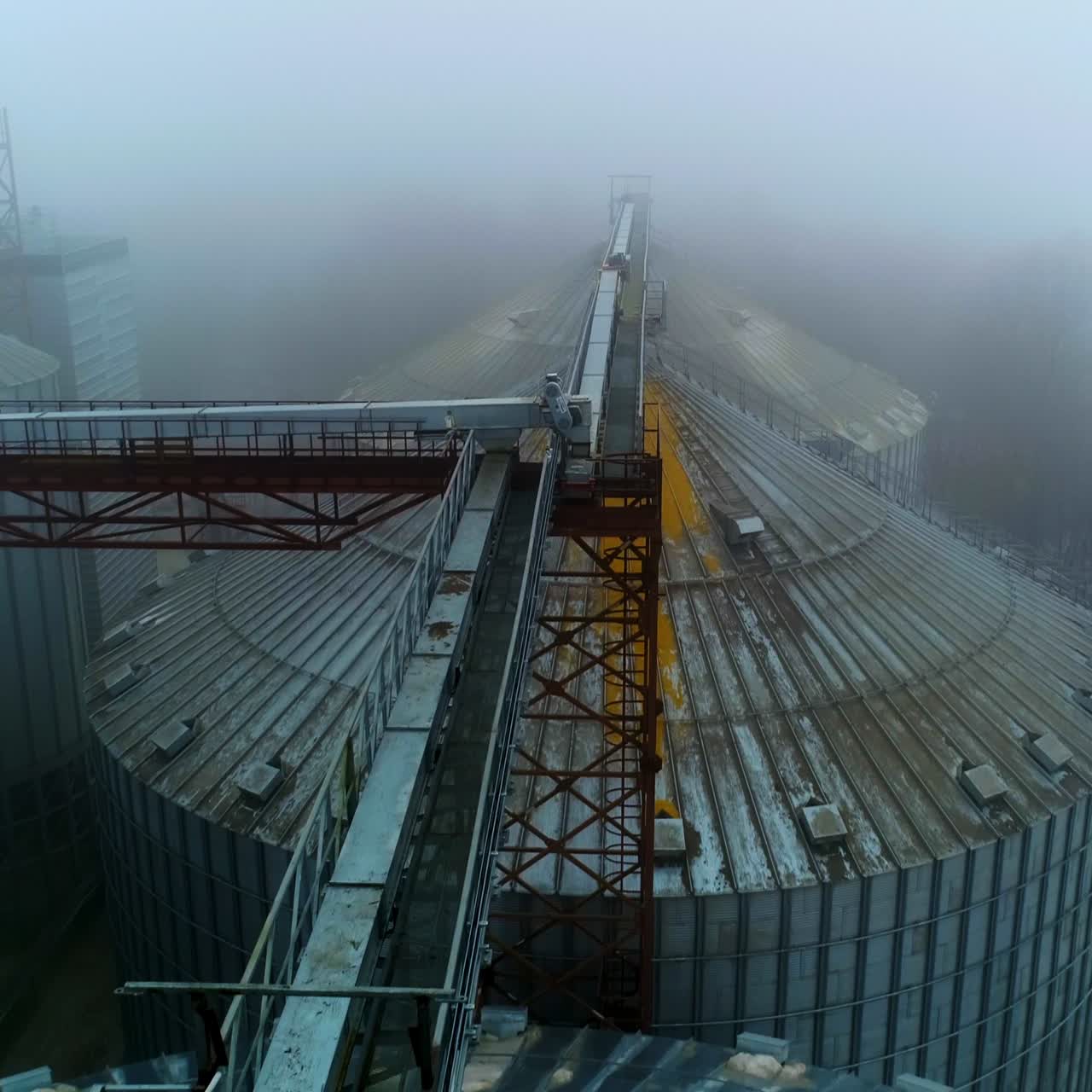 Flying over the tops of metal granary elevators. Huge tanks standing close and connected by supports. Foggy winter day backdrop