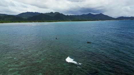 Aerial-shot-of-surfers-in-crystal-waters-off-Agojo-San-Andres,-Catanduanes,-Philippines,-mountains-backdrop