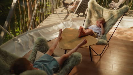 little children rest together on glamping terrace. tired little boy with sister sit on comfortable chairs under clear dome. kids on summer vacation