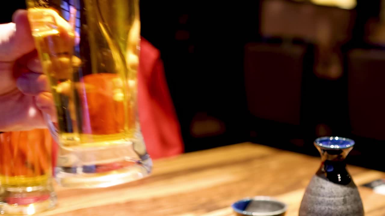 A hand reaches for a frothy beer mug beside a sake set on a wooden table.