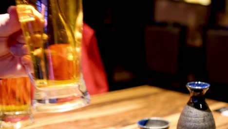 a hand reaches for a frothy beer mug beside a sake set on a wooden table.