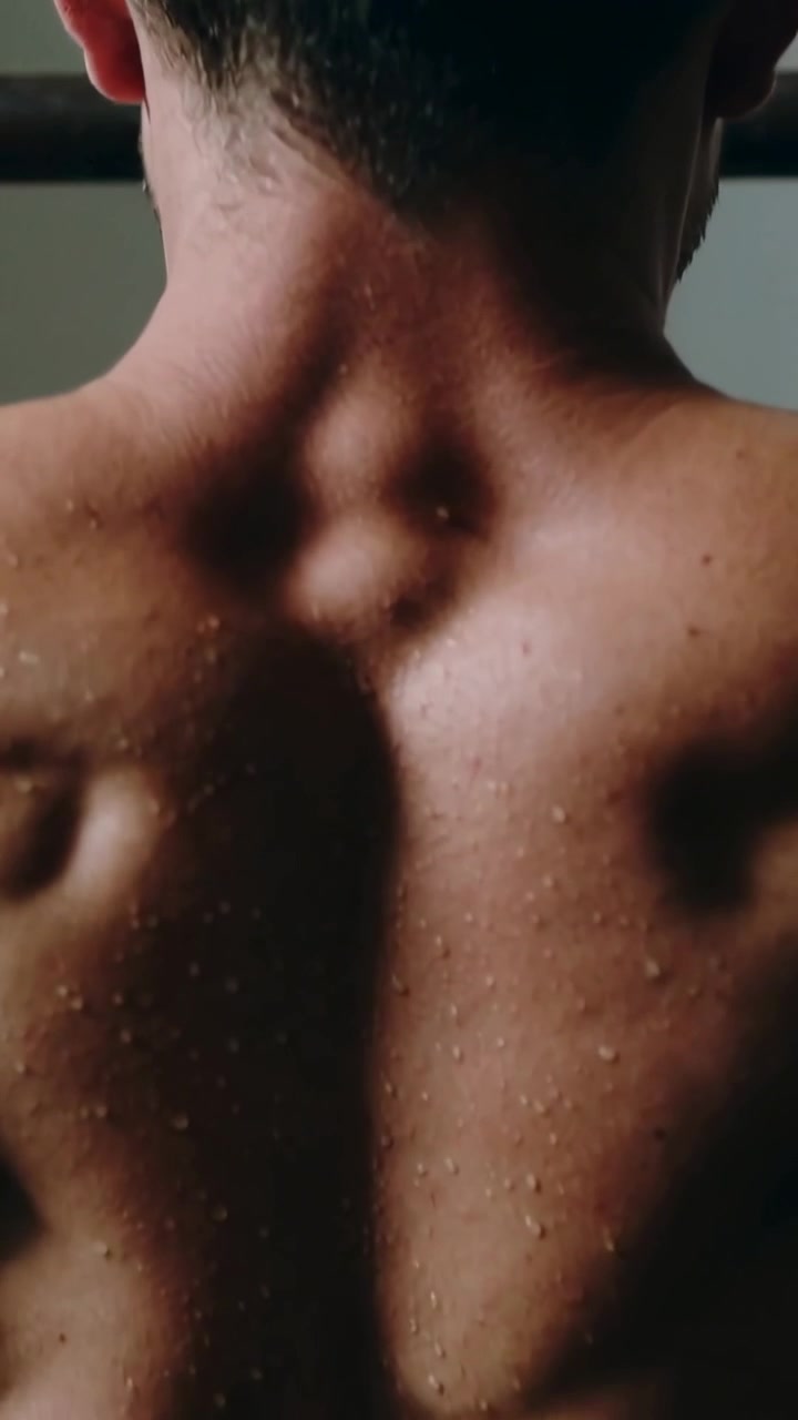 Video verticale: Uomo a torso nudo che afferra la sbarra per trazioni e si tira su, allenandosi nella palestra di casa, con il sudore che luccica.