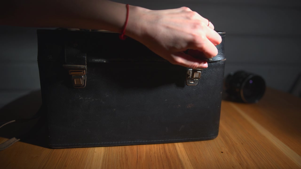 A black leather carrying case with metal latches sits on a wooden table under a narrow warm light beam, with a blurred lens visible in the dark background