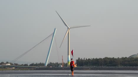 Un-Guardia-Costero-Local-Sosteniendo-Una-Bandera-Roja,-De-Pie-Con-Una-Turbina-Eólica-Gigante-Giratoria-Y-Un-Puente-En-El-Fondo,-área-De-Preservación-De-Humedales-De-Gaomei,-Taichung,-Taiwán