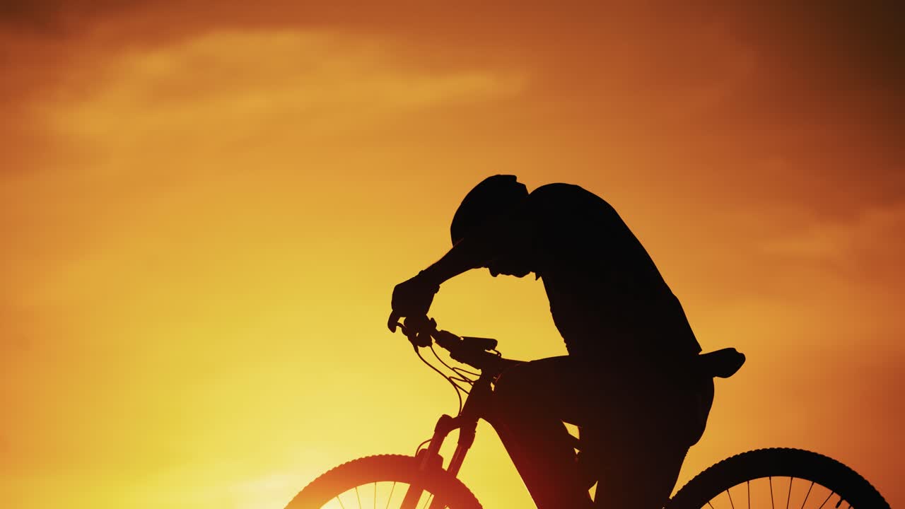 Silhouette of a cyclist man is drinking water after intensive cycling training at sunset. Biker drinks water from bottle and puts it into a bike on the natural golden sun setting background.