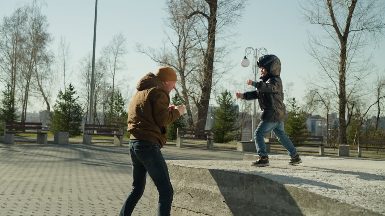 un padre y un hijo jugando, con el hijo avanzando hacia el padre con los puños cerrados, con árboles y postes de luz en el fondo
