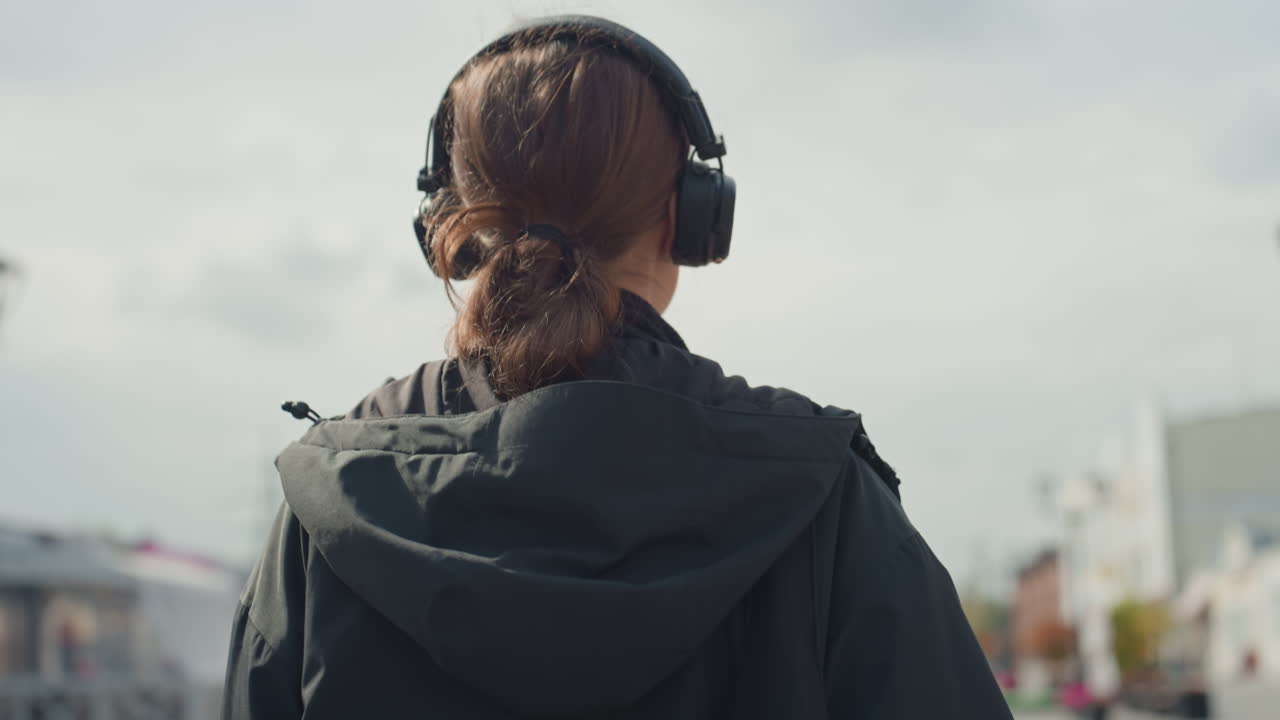 Persona vista da dietro con cuffie, coda di cavallo infilata nella giacca con cappuccio, skyline della città sfocato sotto nuvole soffici, paesaggio sonoro ambientale e andatura costante, immersione nella musica e concentrazione personale, suggerito