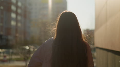 close-up of a girl with long brown hair flowing, wearing a peach jacket and holding a skateboard as she walks through an urban area at sunset. the light casts a warm glow, creating a serene atmosphere