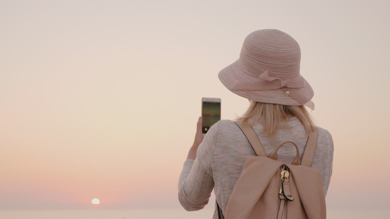 joven con estilo en un sombrero toma fotos del mar y un amanecer rosa en un teléfono inteligente