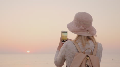 young stylish girl in a hat takes pictures of the sea and a pink sunrise on a smartphone