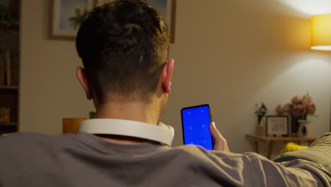 Young Man Wearing Wireless Headphones Spending Evening At Home Sitting On Sofa Looking At Blue Screen Mobile Phone