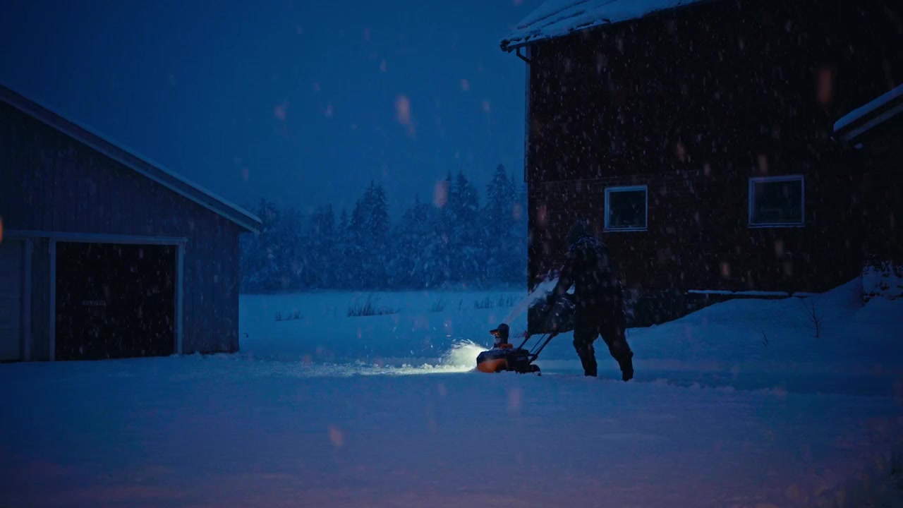 A Person Uses a Snow Blower to Clear Deep Snow Outside Rural Buildings During a Snowy Winter Night - Static Shot