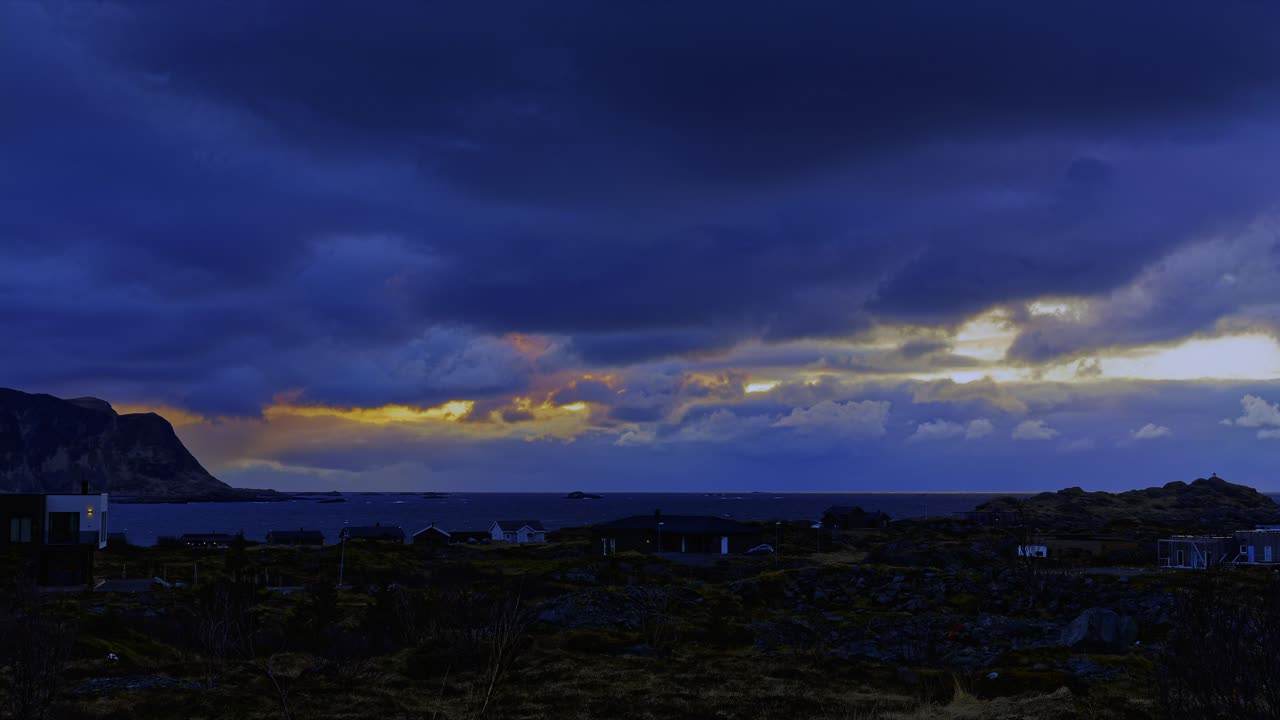 Nordic village by the sea sinks into the evening darkness. Sunset behind heavy clouds above mountain. Time Lapse. Lofoten Islands, Northern Norway.