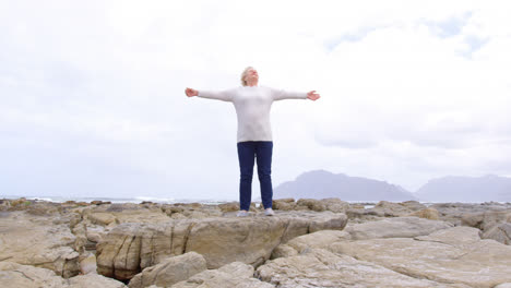 vista frontal de una anciana mujer caucásica de edad avanzada de pie con los brazos extendidos sobre la roca en la playa 4k