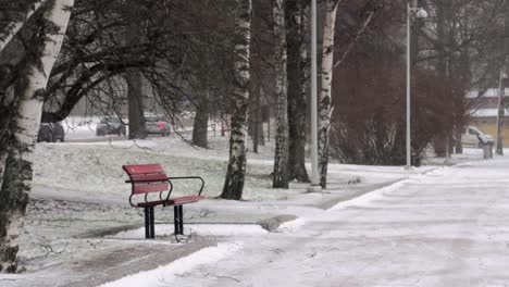 empty bench next to an icy road in a park during a snowstorm, with cars in the distance