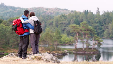 Una-Pareja-De-Adultos-Jóvenes-A-La-Izquierda-De-La-Foto-Parada-En-El-Campo-Admirando-Una-Vista-Al-Lago,-Vista-Posterior,-Lake-District,-Reino-Unido