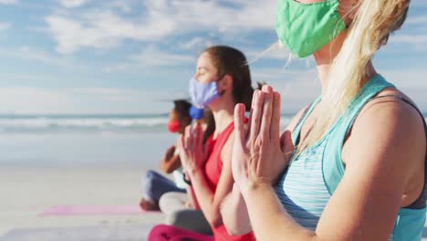 Group-of-diverse-female-friends-wearing-face-masks-meditating-at-the-beach