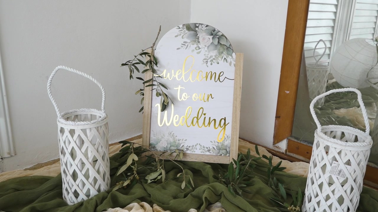 Un cartel de bienvenida para una boda está colocado en un arreglo decorativo. Linternas y hojas verdes rodean el cartel. Este montaje está diseñado para recibir a los invitados a su llegada al lugar.