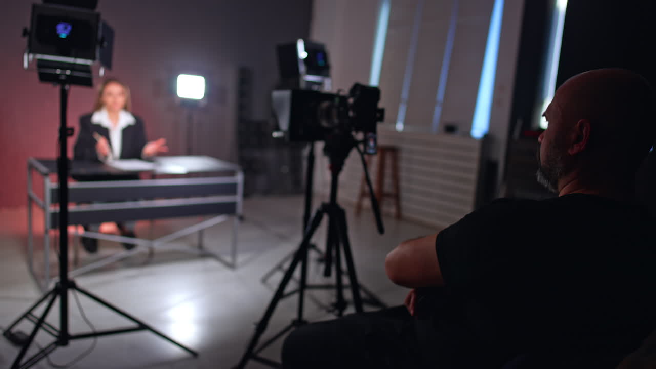 Confident relaxed male photographer sits behind the camera. Woman in black jacket sits at desk speaking for blog.