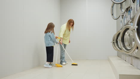 long shot of laundry owner sweeping tiled floor while daughter holds dustpan inside laundromat, teamwork cleaning near industrial washers, family help, responsibility, guidance, routine, hygiene