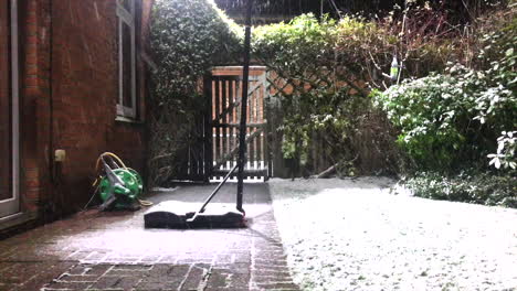 nieve cayendo en el jardín trasero con puerta de madera cerca de la base de un aro de netball y una manguera enrollada
