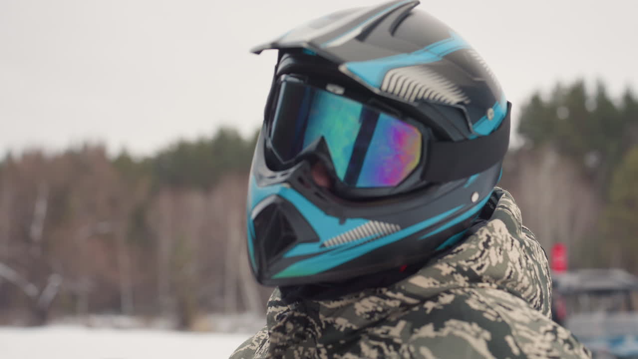 Closeup snowmobile helmet with mirrored goggles, teal and black graphics, visor reflection of winter trees, camo jacket collar, protective padding detail, product style focus on safety gear
