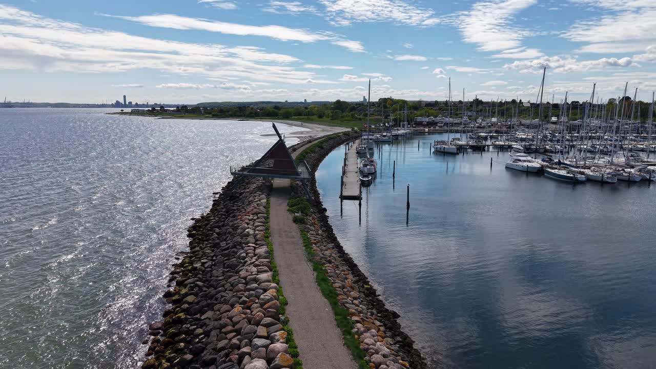 Aerial drone view of a marina harbor with boats docked along the shoreline and a stone jetty stretching into the calm sea