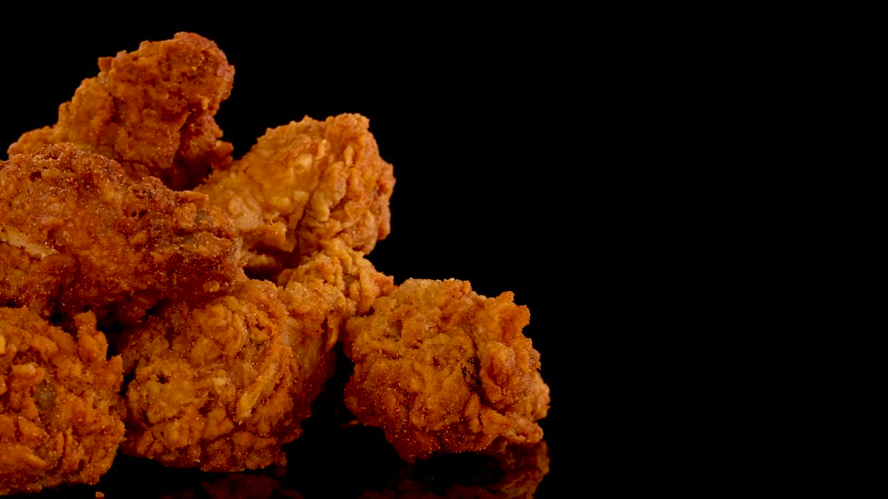Golden, crispy fried chicken pieces are slowly zoomed in on against a black background, highlighting texture, spices, and oily sheen with dramatic studio lighting