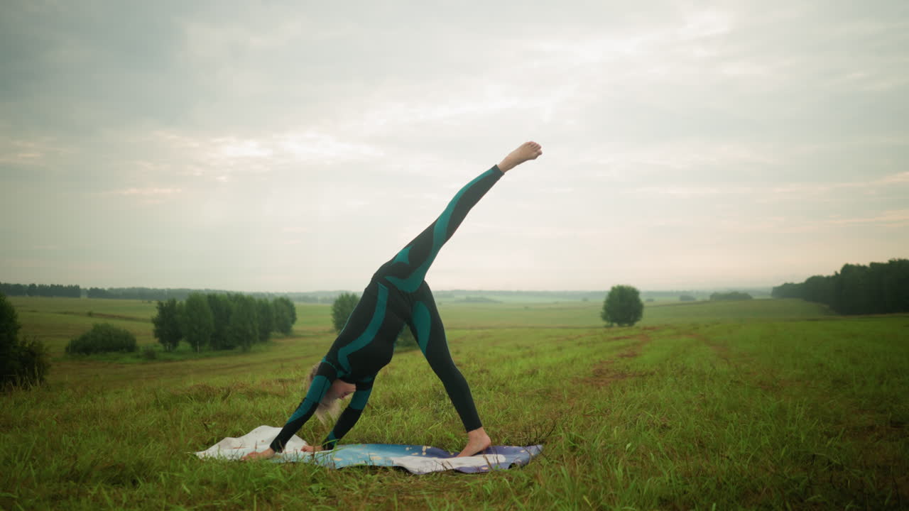 mujer en traje verde y negro practicando yoga dividida de pie al aire libre en una alfombra de yoga en un campo cubierto de hierba bajo un cielo nublado, estirando su cuerpo hacia abajo en un paisaje sereno