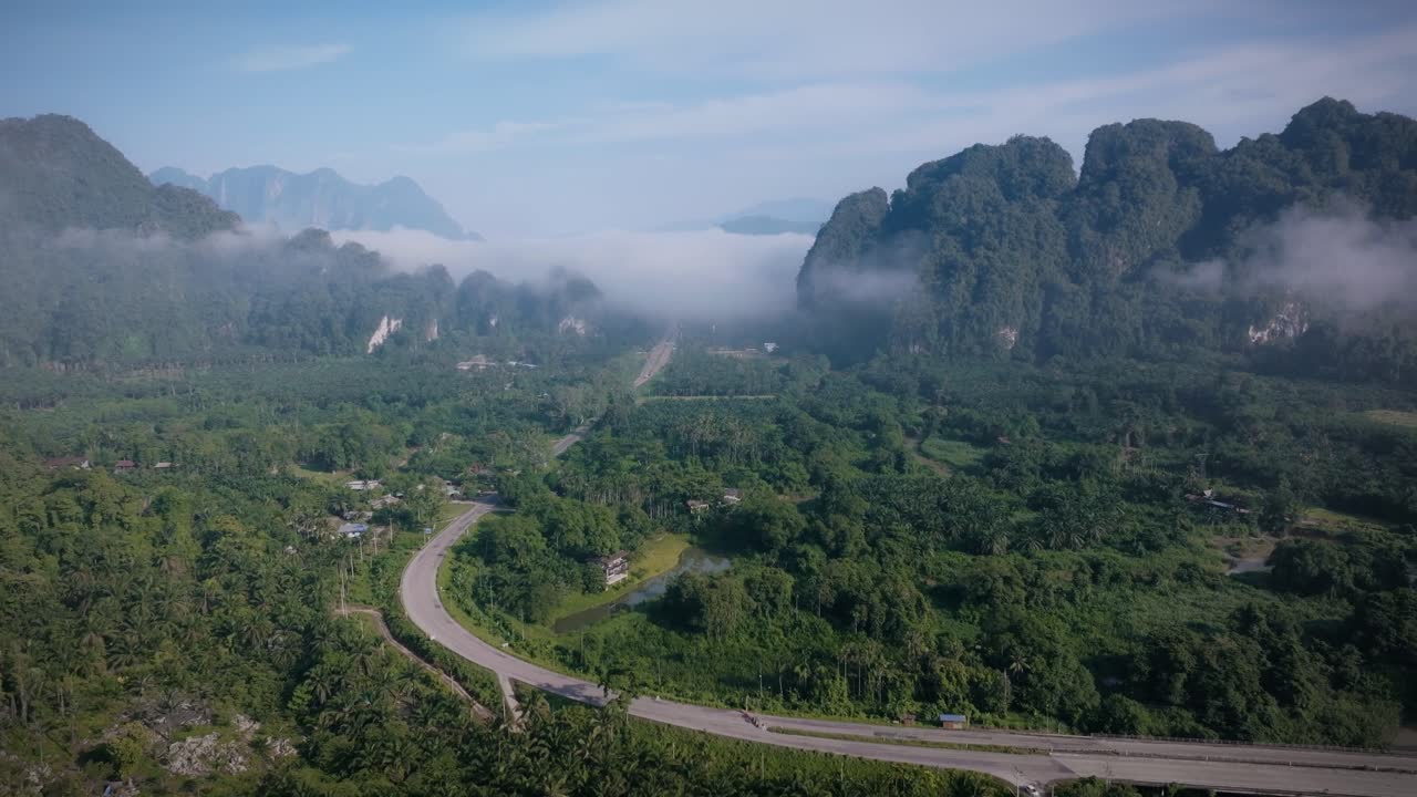 Drone footage of a winding road through the lush green valley of Khao Sok National Park. Morning mist floats over jungle and cliffs, evoking peace and tropical mystery