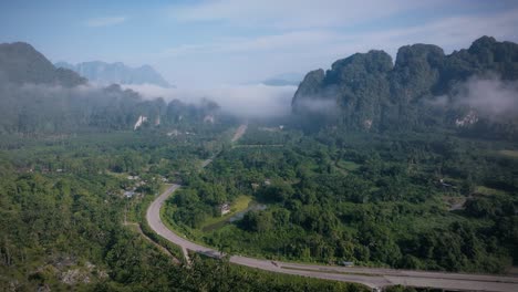drone footage of a winding road through the lush green valley of khao sok national park. morning mist floats over jungle and cliffs, evoking peace and tropical mystery