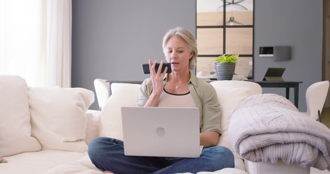 Woman sitting on sofa at home, glancing right, speaking into phone for voice note, using laptop
