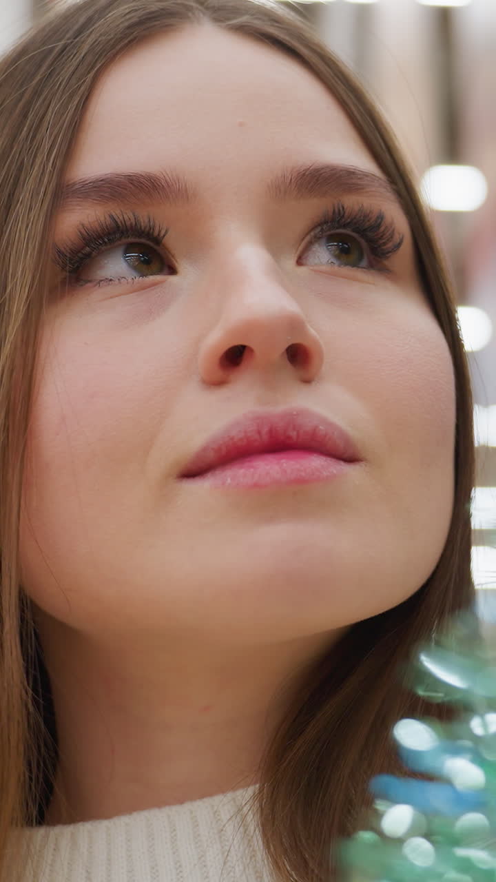Woman with warm expression inspecting green shiny tinsel length in store with bokeh light effect, carefully evaluating decoration options for festive holiday shopping, surrounded by colorful decor
