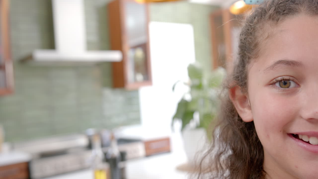 Medio rostro de niña birracial feliz con cabello rizado de pie y sonriendo en una cocina soleada, espacio para texto.
