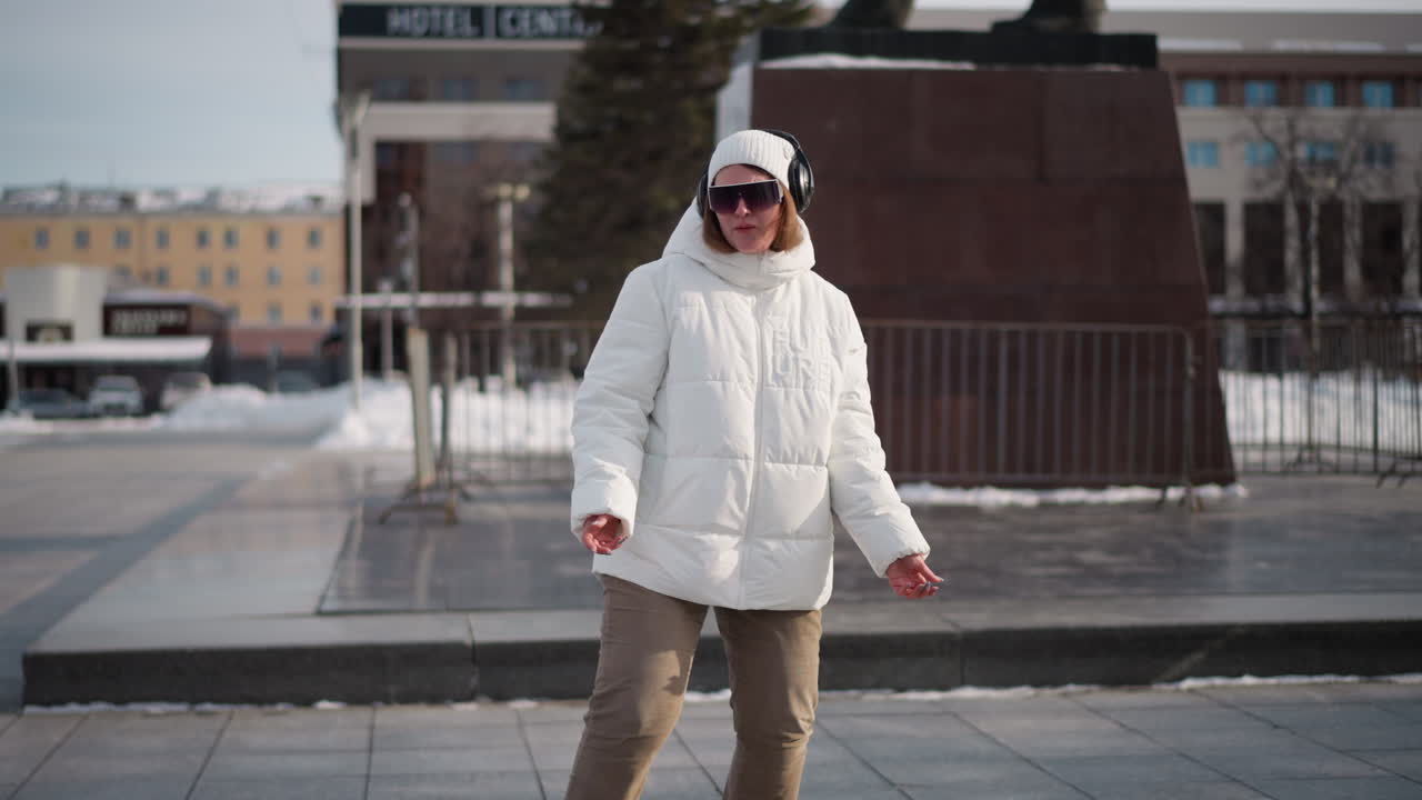 Lady wearing white winter coat, beanie and headset sings and dances expressively with dynamic body and hand movements on wide tiled urban plaza by statue, surrounded by snowy trees in cold season