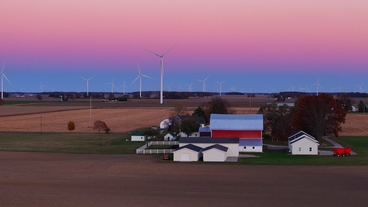 Landbrugsbygninger med dyrket jord. Vindmøller står mod en farverig himmel ved solnedgang.