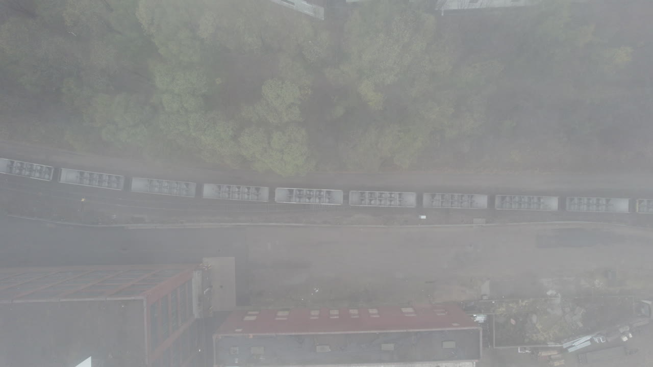 Aerial drone view rising up over a long freight train passing through an industrial area bordered by trees and scattered debris