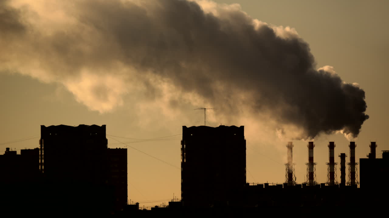 Massive steam cloud rising from multiple industrial chimneys behind dark residential buildings in warm golden backlight Dramatic urban skyline with haze district heating energy infrastructure and emis