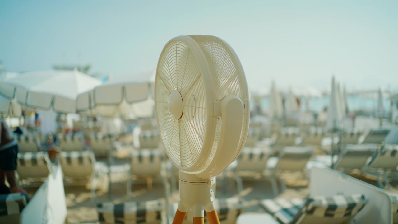 Nærbillede af en hvid elektrisk ventilator på et stativ på en strandklub med liggestole bagved, Cannes, Frankrig.
