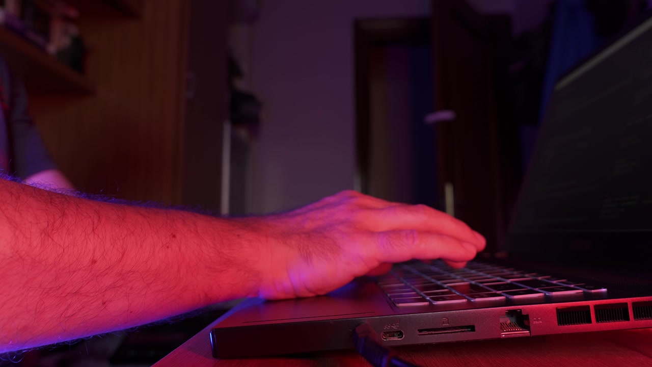 Nærbilde av en persons hender som skriver på et tastatur på en bærbar PC på et hjemmekontor med blå og magenta omgivelsesbelysning. Konsept for programmering, fjernarbeid og teknologi.